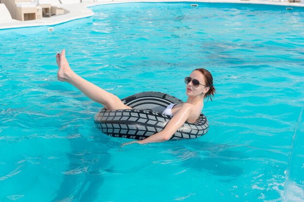 Mujer joven flotando en una piscina azul turquesa relajándose en un tubo de goma con la pierna levantada en el aire mientras disfruta de sus vacaciones de verano