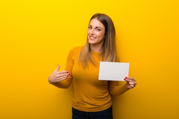 La mujer joven en el fondo amarillo que lleva a cabo un cartel vacío para inserta un concepto