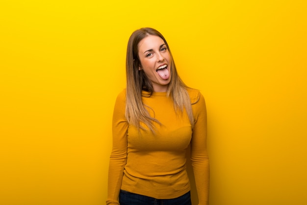 Mujer joven en el fondo amarillo que muestra la lengua en la cámara que tiene mirada divertida