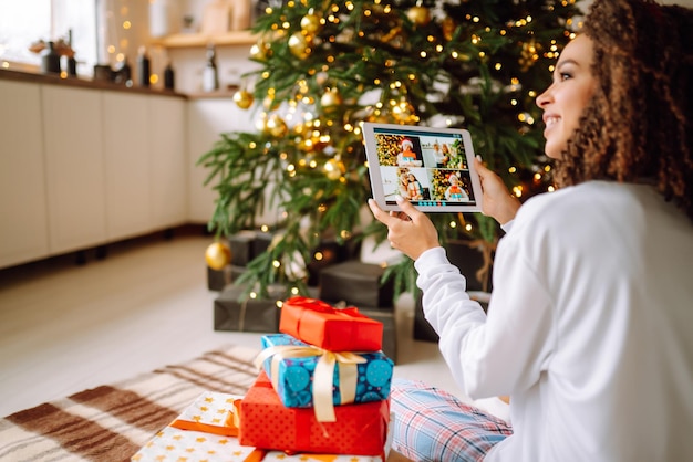 Una mujer joven en el fondo del árbol de Navidad con regalos con tableta tiene videollamada o videochat