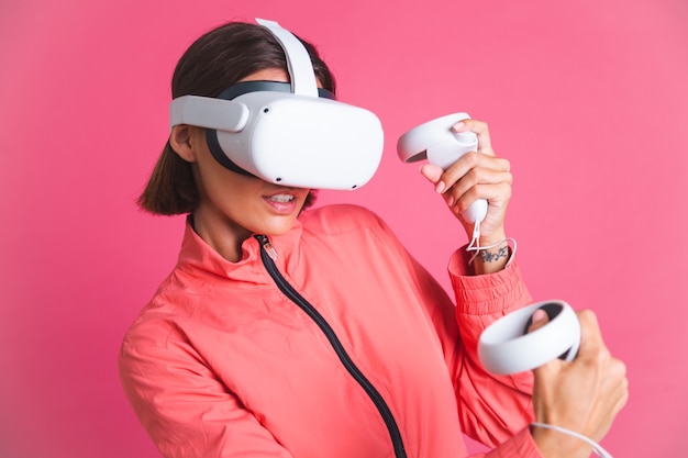 Mujer joven en forma con chaqueta deportiva y gafas de realidad virtual jugando juegos de lucha de boxeo en rosa