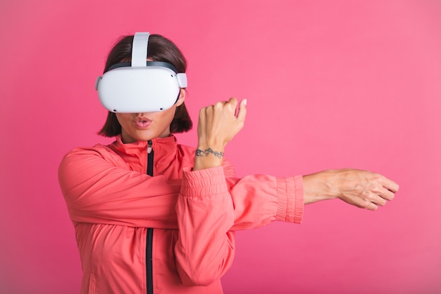 Mujer joven en forma con chaqueta deportiva y gafas de realidad virtual que se estira en rosa