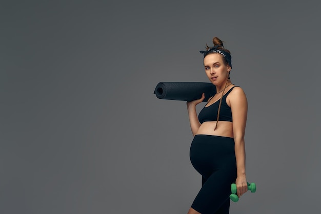 Mujer joven en forma haciendo ejercicio durante su embarazo de pie sonriendo a la cámara con agua embotellada en una mano y una colchoneta de gimnasia en la otra