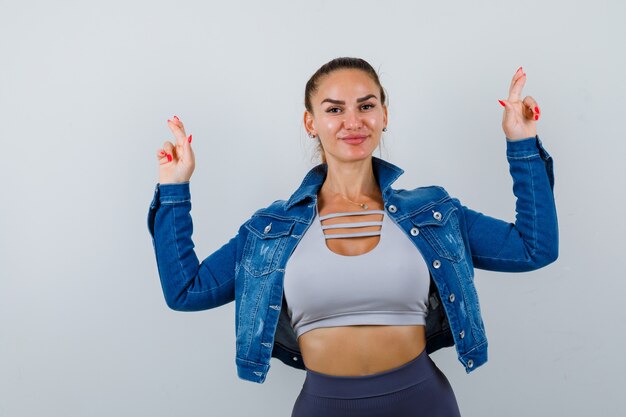 Mujer joven en forma en la parte superior, chaqueta de mezclilla que muestra los dedos cruzados y se ve alegre, vista frontal.