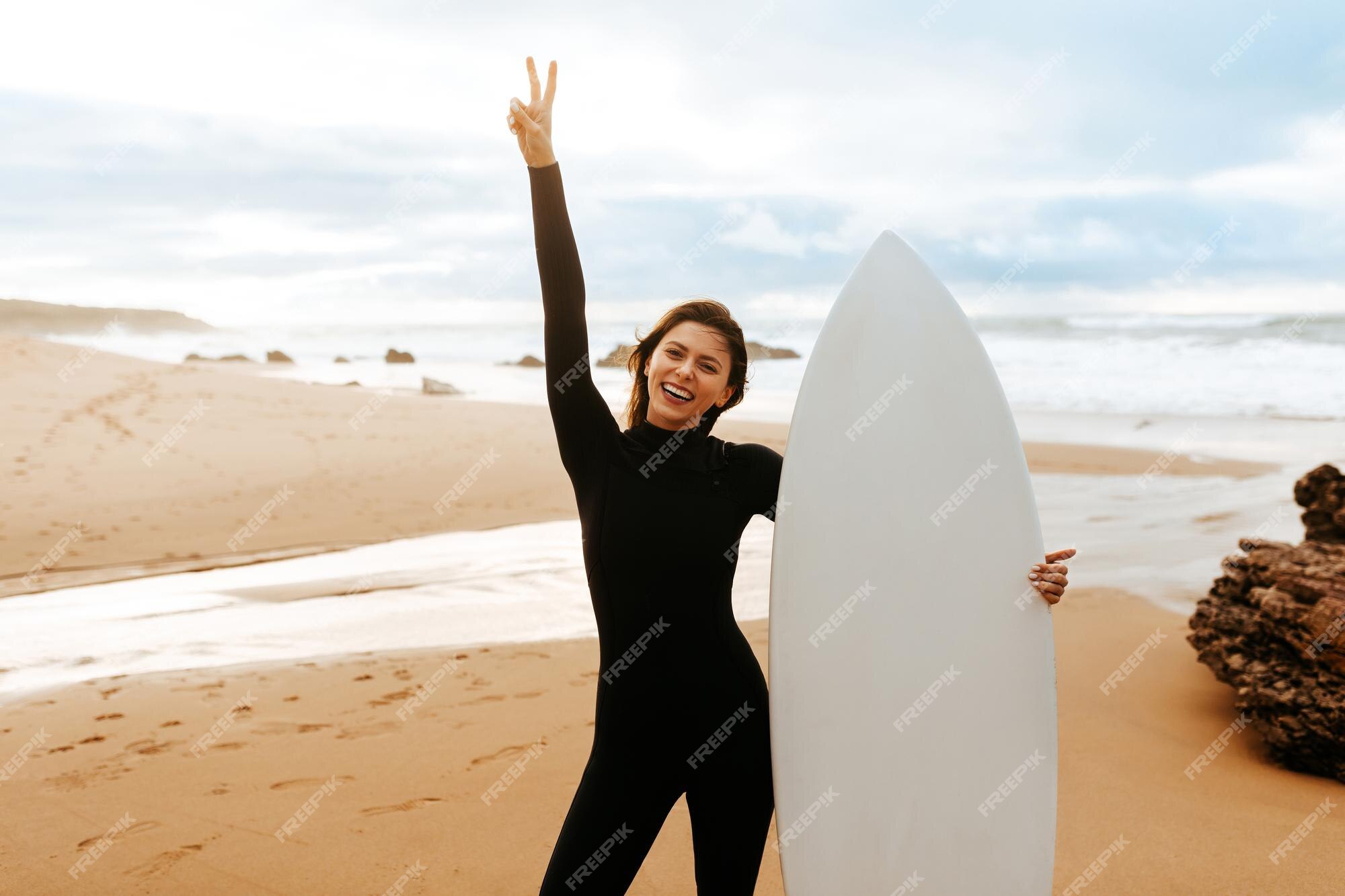 Mujer joven en forma de pie con tabla de surf en la playa junto al mar sonriendo a la cámara y gesticulando signo de la paz | Foto Premium