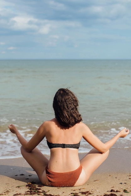 Mujer joven en forma practicando yoga en la playa sentada en la arena y mirando las olas del mar chica meditando en vacaciones de verano concepto de salud mental y autocuidado