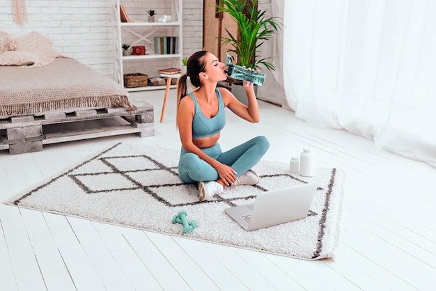 Mujer joven en forma en ropa deportiva bebe agua mientras hace ejercicios de calentamiento todos los días por la mañana en el piso