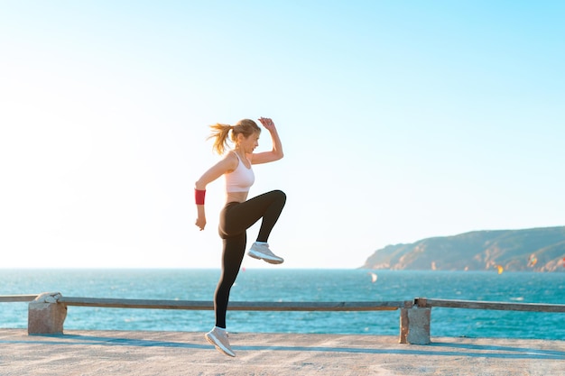 Mujer joven en forma saltando alto en la playa con entrenamiento femenino adulto joven caucásico activo al atardecer con ejercicio de entrenamiento de peso corporal para la automotivación de la mujer