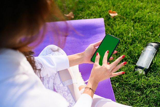 Mujer joven en forma sentada en la estera de deporte púrpura en el parque, sosteniendo el teléfono inteligente y agrandar la pantalla durante el entrenamiento, fitness al aire libre. Smartphone móvil con pantalla verde.