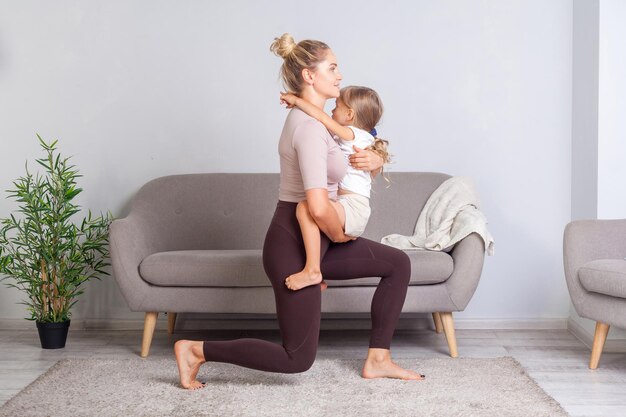 Mujer joven en forma sosteniendo a su hija preescolar en sus brazos y en cuclillas sobre una rodilla familia feliz haciendo ejercicio de gimnasia juntos en casa entrenamiento físico con cuidado del cuerpo infantil estilo de vida saludable