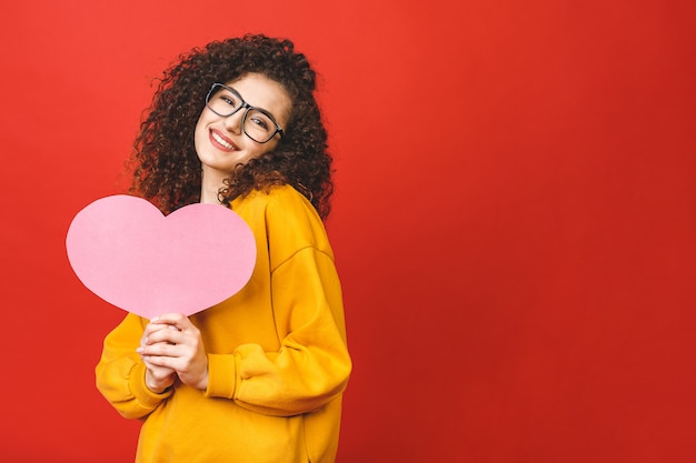 Mujer joven de la foto que lleva a cabo el corazón grande del papel rosado sobre fondo del color rojo.