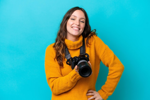 Mujer joven fotógrafa aislada de fondo azul posando con los brazos en la cadera y sonriendo