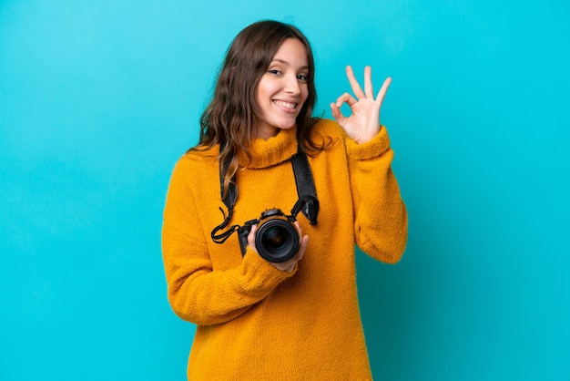 Mujer joven fotógrafa aislada sobre fondo azul que muestra el signo ok con los dedos