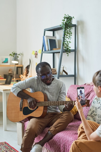 Mujer joven fotografiando a joven en el teléfono móvil mientras toca la guitarra en casa