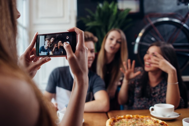 Mujer joven fotografiando a sus amigos positivos en la cafetería usando el fondo del teléfono inteligente desenfocado