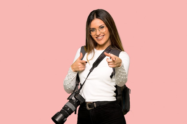 Mujer joven fotógrafo apuntando hacia el frente y sonriendo en pared rosa aislado