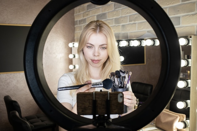 Una mujer joven se para frente a una lámpara de anillo y realiza cursos en línea sobre cómo aplicar el maquillaje.
