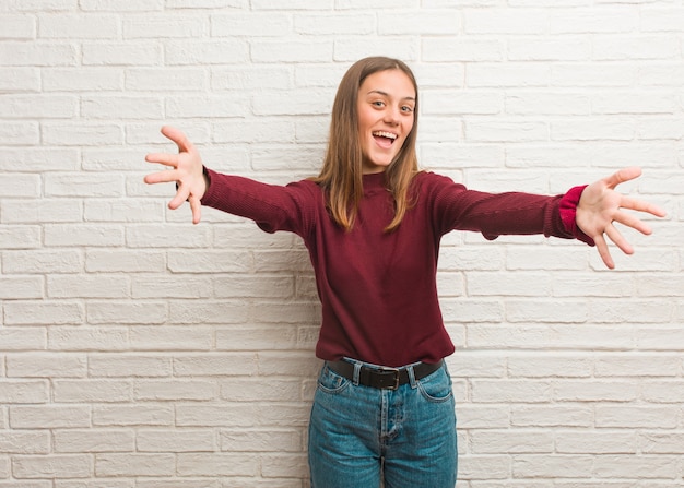 Mujer joven fresca sobre una pared de ladrillos muy feliz dando un abrazo al frente