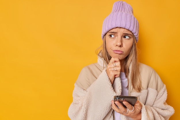 Mujer joven frustrada molesta sostiene la barbilla mira hacia otro lado mensajes de tipo en el teléfono celular lee malas noticias en las redes sociales usa gorro de punto y chaqueta aislada sobre fondo amarillo vivo espacio de copia en blanco