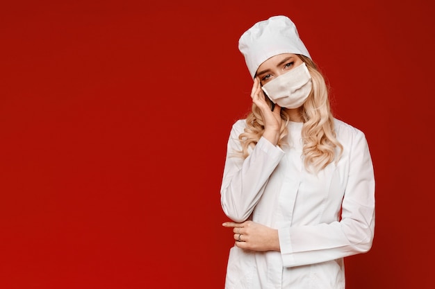 Mujer joven frustrada en uniforme médico y máscara protectora que siente dolor de cabeza y que mira in camera, aislado en el fondo rojo, copia el espacio. Concepto de salud.