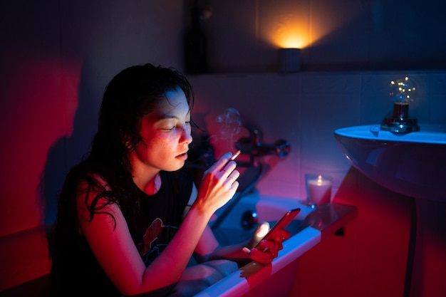 Mujer joven fumando cigarrillos desplazándose por las redes sociales en el teléfono inteligente mientras está sentada en la bañera