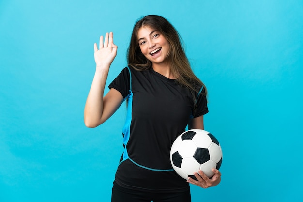 Mujer joven futbolista aislada en azul saludando con la mano con expresión feliz