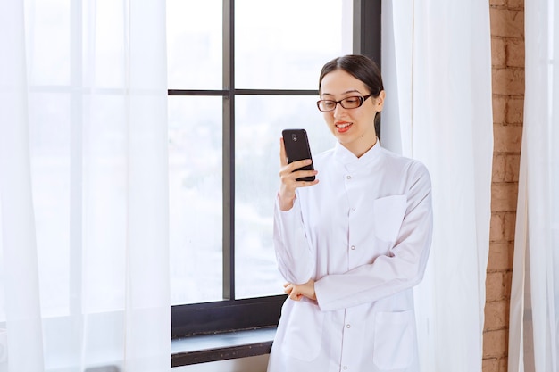 Mujer joven con gafas en bata de laboratorio sosteniendo la comprobación de mensajes en el teléfono móvil junto a la ventana.