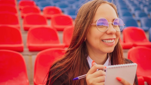 Mujer joven en gafas con bolígrafo sentado en las gradas del estadio sola Periodista femenina escribiendo notas durante las competiciones en el estadio de la calle