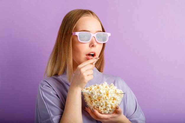 Mujer joven en gafas de cine viendo películas en 3D en el cine. Sonriente espectador de películas de chica adolescente en vasos comiendo palomitas de maíz aislado sobre la pared de color púrpura con espacio de copia.