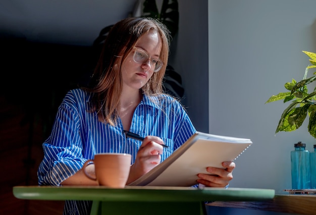 Mujer joven en gafas escribiendo con bolígrafo en el cuaderno tomando notas en el café con taza de café en la mesa