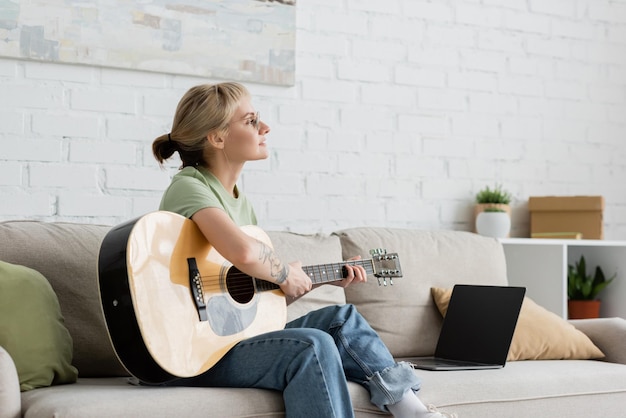mujer joven con gafas con flechazo y tatuaje tocando la guitarra acústica cerca de la computadora portátil con pantalla en blanco