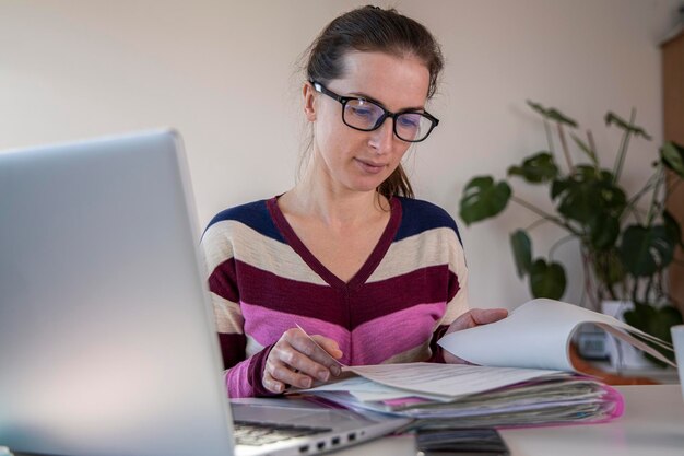 Foto mujer joven con gafas mirando papeles mientras está sentada frente a una computadora portátil