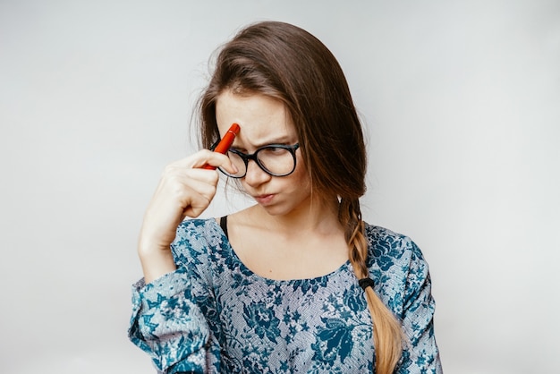 Una mujer joven con gafas pensó en problemas, frunció el ceño, sostiene un bolígrafo en la mano, se frota la frente