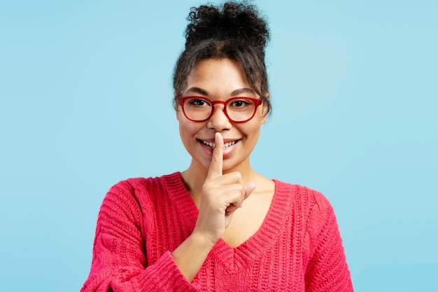 Mujer joven con gafas rojas haciendo un gesto de silencio sobre un fondo azul