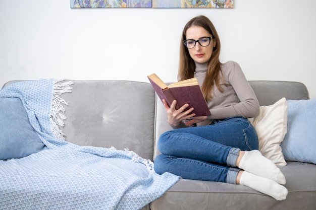 Mujer joven con gafas sentada en un sofá leyendo un libro