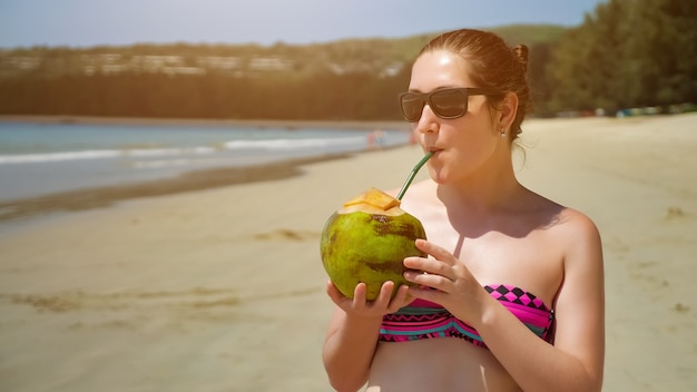 Mujer joven en gafas de sol bebe coco en la playa copyspace