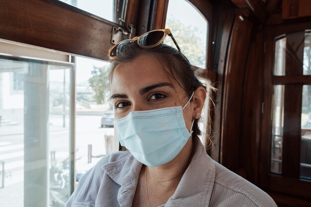 Mujer joven con gafas de sol y una mascarilla dentro de un viejo tren en oporto sonriendo a la cámara en concepto de ropa indie, viajes y nuevas experiencias