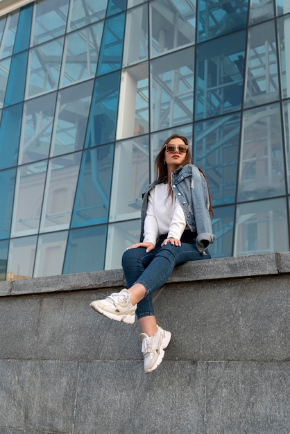 Mujer joven en gafas de sol en ropa juvenil en la calle de la ciudad Atractiva mujer elegante descansando en la calle de la ciudadVertical