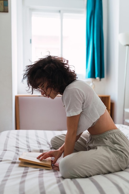 Mujer joven con gafas de sol sentada en su cama leyendo un libro