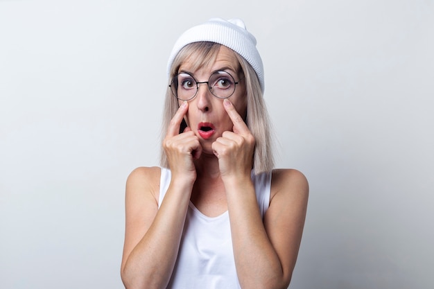 Mujer joven con gafas sosteniendo sus dedos cerca del ojo sobre un fondo claro.