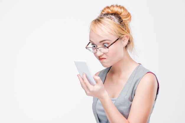 Mujer joven con gafas con teléfono móvil se ve enojada, molesta e irritada. Rubia molesta y gruñona con smartphone en pared gris con espacio de copia