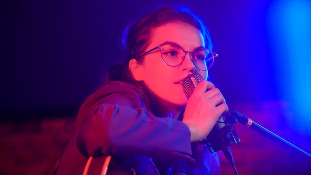 Una mujer joven con gafas tocando la guitarra y cantando con luces de neón