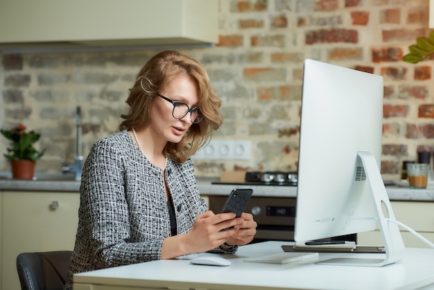 Una mujer joven con gafas trabaja remotamente en una computadora de escritorio en su estudio. Una jefa usa un teléfono inteligente durante una videoconferencia en casa.