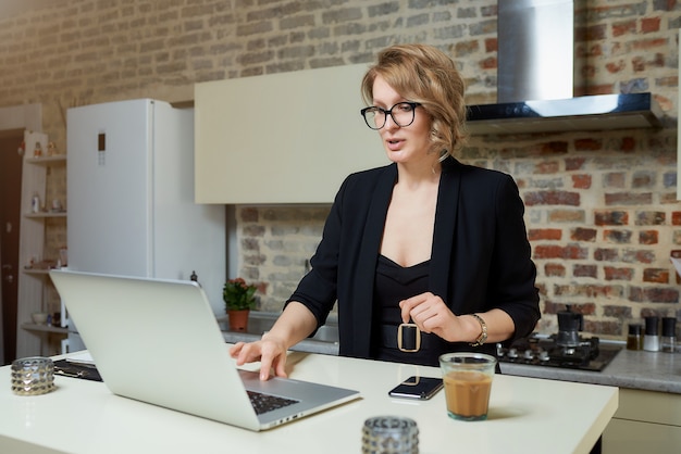 Una mujer joven con gafas trabaja remotamente en una computadora portátil en su cocina. Una chica seria navegando por las noticias en internet en casa ...