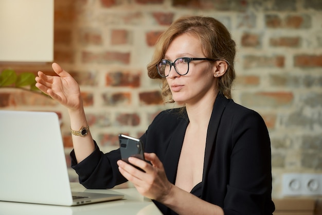 Una mujer joven con gafas trabajando remotamente con un teléfono inteligente en un portátil. Una mujer rubia discutiendo un problema con colegas en una video conferencia en casa.