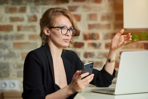 Una mujer joven con gafas trabajando remotamente con un teléfono inteligente en un portátil. Una mujer rubia discutiendo un problema con colegas en una video conferencia en casa.