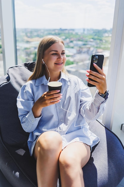 Mujer joven gerente con auriculares hablando por teléfono y tomando café mientras está sentada en un espacio de coworking moderno Chica independiente que trabaja de forma remota en línea