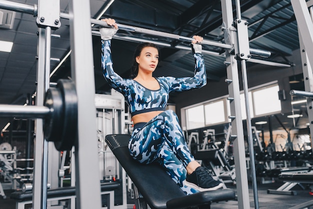 Mujer joven en el gimnasio con aparatos de gimnasia. Hermosa mujer delgada trabajando duro con barra. Ella está mirando hacia abajo porque está demasiado concentrada en el proceso. De cerca