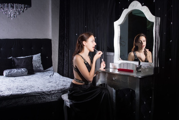 Mujer joven glamorosa aplicar maquillaje frente al espejo en un elegante dormitorio en la sombra de la luz