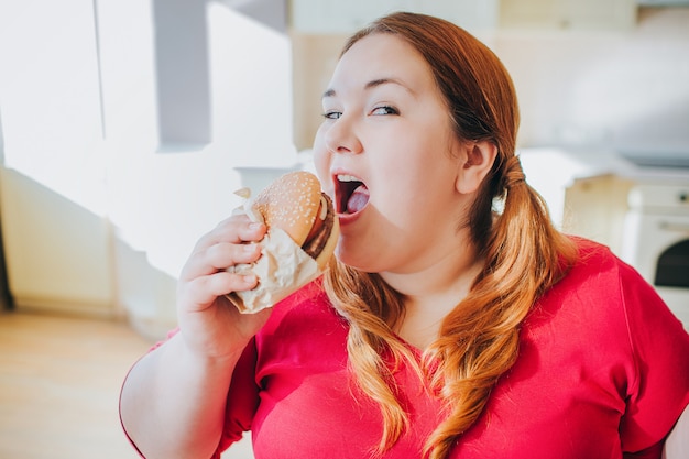 Mujer joven gorda en la cocina que se sienta y que come la comida basura.